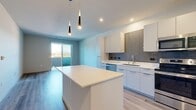 This thoughtfully designed single-floor residence offers a clean, contemporary aesthetic throughout its well-organized layout. The open-concept kitchen serves as the centerpiece of the living area, showcasing sleek white cabinetry, a gray subway tile backsplash, white quartz countertops, and a full suite of stainless steel appliances including a refrigerator, range, microwave, and dishwasher. A white center island provides additional prep space and casual seating, while stylish pendant lights hang above. Light wood-style flooring flows throughout the main living areas, enhancing the bright and airy atmosphere. Sliding glass doors open to a private balcony, framing views of the surrounding landscape and mountains beyond. The spacious primary bedroom is finished with plush carpeting and features a large window that captures natural light and scenic views. A dedicated walk-in closet with built-in shelving and hanging rods provides ample storage. The bathroom is clean and functional, offering a full soaking tub and shower combination, built-in wall shelving, and wood-style flooring that ties the space together. A hallway connects the private areas of the unit to the main living space, completing a practical and cohesive floor plan.