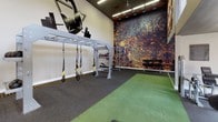 Located at 3910 Mesa Dr in Oceanside, California, this single-story fitness facility delivers a thoughtfully designed workout environment suited for a wide range of training styles. The open floor plan maximizes usable space, with distinct zones dedicated to both strength training and functional movement.

The main workout area features rubber flooring throughout, transitioning to a turf surface that defines the functional training zone — ideal for sled work, agility drills, and dynamic exercises. A full-wall mirror runs along one side, visually expanding the space and allowing users to monitor their form during workouts.

Strength equipment is well-distributed across the floor, including a power rack with an adjustable bench, a multi-use functional training rig equipped with TRX suspension straps, and a rack stocked with medicine balls and kettlebells. A dumbbell tower provides easy access to free weights of varying sizes.

The facility's interior design adds considerable visual energy. Bold graphic murals — including a dramatic abstract textured wall in deep rust and gold tones — contrast with clean white walls adorned with athletic-themed artwork. Orange diamond-patterned accent panels near the ceiling add a dynamic, branded aesthetic. Overhead linear lighting ensures consistent illumination throughout. A wall-mounted flat-screen television provides entertainment during training sessions, and an electrical panel and entryway are accessible near the facility's entrance.