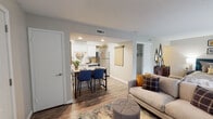 This single-floor unit is designed with an open, functional layout that connects the main living spaces seamlessly. The living area features wood-look flooring, neutral walls, and a textured ceiling, creating a warm and inviting atmosphere. A sofa and ottoman anchor the space, which flows directly into the sleeping area — accommodating both a bed and living furniture within the same open zone. A fireplace with a tile surround is visible along one wall, and a sliding glass door provides access to an outdoor area while drawing in natural light.

The kitchen is accessible via an open pass-through and features white shaker-style cabinetry, quartz countertops, a subway tile backsplash, and stainless steel appliances including a refrigerator, range, and microwave. A bar-height counter with seating for two serves as the primary dining area. The kitchen is compact yet well-organized, offering ample cabinet storage.

The bathroom includes a wide vanity with white cabinetry, quartz countertops, an undermount sink, and a large wall mirror that reflects into the adjacent bedroom. A walk-in closet is accessible from the main living area, providing dedicated storage. The unit's cohesive finishes and efficient use of space make it a well-appointed single-floor residence.