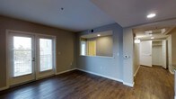 This single-floor residence offers a thoughtfully designed layout that balances comfort and functionality. The living area serves as the central focal point, showcasing rich, dark hardwood flooring that runs throughout and provides a warm, grounded aesthetic. Neutral gray and taupe wall tones complement the clean white baseboards and ceiling, creating a modern and cohesive interior palette.

A defining architectural feature is the pass-through window cutout between the living area and the kitchen, promoting an open and connected feel between the two spaces. This design element allows natural light from the kitchen's window to filter into the living area, enhancing the overall brightness of the unit. A door with slatted blinds on the exterior-facing wall admits additional natural light and offers convenient access to the outdoors.

Practical details are well-integrated throughout, including ceiling-mounted HVAC vents, a wall-mounted thermostat, and recessed lighting within the pass-through nook. A hallway visible toward the rear of the space suggests access to additional rooms, further extending the functional layout. Overall, the space presents a clean, move-in-ready environment with durable finishes and a well-considered floor plan suited to everyday living.