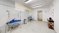 Located in Hooksett, New Hampshire, this dedicated dog spa is a compact, single-room facility thoughtfully designed to accommodate professional pet grooming services. The space features a speckled terrazzo-style floor that is both durable and easy to clean, complemented by bright white walls adorned with playful dog-themed decals — including paw prints and bone motifs — that lend a welcoming atmosphere.

Overhead fluorescent lighting ensures the room is well-illuminated throughout, supporting precise grooming work. A wall-mounted climate control unit helps maintain a comfortable environment for both pets and staff year-round. The centerpiece of the grooming area is a stainless steel professional bathing station, equipped with anti-slip floor grates for safety. A grooming table with an adjustable restraint arm provides a dedicated space for drying and styling.

A wooden multi-tier shelving unit offers organized storage for grooming supplies and pet bowls, while a stainless steel step-pedal waste bin supports cleanliness and hygiene. A black metal bench provides seating, likely for waiting pet owners. The room is accessed through a single doorway fitted with a welcome mat. Fire safety is addressed with a wall-mounted alarm. Overall, the space is efficiently laid out, clean, and purpose-built to deliver professional dog grooming services in a safe and organized environment.