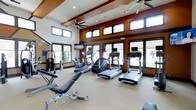 This single-floor fitness center offers a comprehensive workout environment designed to accommodate a variety of exercise routines. The space is anchored by a row of modern cardio machines, including multiple treadmills and a stair climber, all positioned to take advantage of the expansive floor-to-ceiling windows that line the walls and flood the room with natural light. A dedicated strength training area features a multi-station cable machine, adjustable weight bench, and free weights, providing everything needed for a full resistance workout. The soaring vaulted ceiling, accented by warm earth-toned wall panels and exposed structural beams, lends the space an open, airy feel. Dual ceiling fans are mounted overhead to ensure comfortable airflow throughout workout sessions. Recessed lighting supplements the abundant natural light from the dual rows of clerestory and large lower windows, keeping the space bright at any time of day. A wall-mounted flat-screen television is also on hand for entertainment during cardio sessions. The overall design balances functionality with an inviting aesthetic, making this fitness center a well-appointed destination for daily exercise.