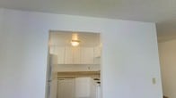 This single-floor residence offers a straightforward and functional layout designed for comfortable everyday living. The space is defined by its clean, neutral palette, with white walls and a textured popcorn ceiling that extends throughout, creating a cohesive and understated aesthetic.

The kitchen is accessed through an arched pass-through opening, giving it a semi-open connection to the adjacent living area. Inside, the kitchen features white upper and lower cabinetry that provides ample storage, complemented by a light-toned countertop. Essential appliances are included, among them a refrigerator, dishwasher, and a stovetop range, all finished in white to match the cabinetry for a uniform look. A flush-mount ceiling light illuminates the kitchen, ensuring the space remains bright and practical.

The living area, visible from the kitchen pass-through, continues the same white-wall finish and textured ceiling treatment, contributing to an open and airy feel. The simple, uncluttered design of the space lends itself well to a variety of furnishing styles and personal touches. Overall, this residence presents a clean, move-in-ready environment with a functional design suited for efficient, day-to-day living.