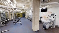 This fitness center offers a comprehensive workout environment designed to accommodate a wide range of exercise routines. The space features an open, industrial aesthetic with exposed ceiling infrastructure, including visible ductwork and piping painted white, which contributes to the room's airy, loft-like character. Concrete structural columns punctuate the floor plan, adding to the raw architectural appeal.

The cardio zone is lined with a row of treadmills, providing ample options for endurance training. Nearby, a stationary bike and exercise balls offer additional low-impact workout choices. On the opposite side of the space, a dedicated strength training area features a rack of free weights, adjustable benches, and cable machines positioned against a bold olive-green accent wall with a mirror, creating an energizing atmosphere for resistance workouts.

A large motivational typographic mural dominates one wall, featuring fitness-related keywords in a vibrant, multi-colored design that adds personality and energy to the space. Overhead, a mounted flat-screen television provides entertainment during workouts. The flooring throughout is a durable blue-gray rubberized surface, practical for heavy equipment and high-traffic use. The space is well-lit with a combination of fluorescent and pendant lighting, ensuring excellent visibility across all workout zones.