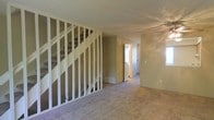 This two-story space presents a functional layout with a carpeted main living area that extends toward a hallway leading to additional rooms. A staircase with an open baluster frame serves as a central architectural element, connecting the two floors and adding visual structure to the space. The open design of the stair railing allows light to pass through, contributing to a relatively airy feel in the main level.

The living area is finished with neutral-toned carpet and beige walls, providing a blank canvas suitable for a range of interior styles. A ceiling fan is installed overhead, enhancing air circulation throughout the space. Natural light enters through a window visible along the far wall, brightening the interior.

The hallway visible beyond the main area leads to what appears to be additional rooms, including a bathroom, suggesting a practical multi-room layout on the main floor. Floor vents indicate a central HVAC system is in place. Some areas of the space appear to be mid-renovation, with exposed framing visible, indicating opportunities for finishing or customization to suit specific needs.