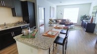 This single-floor residence is designed around an open-concept layout that connects the kitchen, dining area, and living room into one cohesive, functional space. The kitchen is outfitted with rich, dark espresso cabinetry that provides a bold contrast against the light-toned granite countertops. The expansive kitchen island serves as both a food preparation surface and an informal dining area, comfortably accommodating bar-style seating with sleek black stools — making it ideal for casual meals or entertaining. The kitchen also features a full suite of appliances integrated seamlessly into the cabinetry.

Wood-style flooring extends throughout the main living space, adding warmth and visual continuity. The living area is open and airy, benefiting from a large window that allows abundant natural light to fill the room. The layout creates a natural flow between the kitchen and living zone, making the space feel larger and well-connected.

A doorway visible from the main area leads to a separate bedroom, providing a private retreat while maintaining the open feel of the common spaces. Overall, the design balances practicality with modern aesthetics through clean lines, quality finishes, and thoughtful spatial planning.