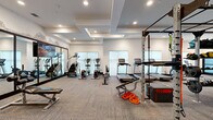 This well-appointed fitness center offers a comprehensive workout environment across a single, expansive floor. The space is designed with a modern aesthetic, featuring a coffered ceiling with recessed lighting, neutral-toned herringbone-patterned carpet, and crisp white walls accented by exposed brick detailing that adds a stylish industrial touch.

The cardio zone is well-equipped with multiple treadmills, elliptical trainers, stair climbers, and stationary bikes, all positioned near large windows that allow ample natural light to fill the space. A full-length mirror wall in this section enhances the sense of openness and allows users to monitor their form.

The strength and functional training area is equally impressive, featuring a multi-station cable and power rack system, a full dumbbell rack, adjustable benches, medicine balls, battle ropes, a mini trampoline, and suspension training equipment. This zone supports a wide range of functional fitness and resistance training workouts.

A glass-partitioned room with black steel-framed doors provides a dedicated area for additional equipment or private training sessions. Wall-mounted flat-screen televisions are positioned throughout the space for entertainment. The overall layout is thoughtfully organized to maximize usability, offering a professional-grade fitness experience in a clean, modern environment.