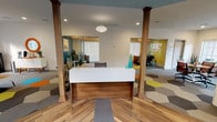This single-story commercial office space presents a thoughtfully designed interior that balances open collaboration with private work areas. The central reception area anchors the layout, featuring a sleek two-tone desk with a white laminate top and warm walnut base, framed by decorative wood columns that extend throughout the space. The flooring transitions seamlessly between wood-look planks near the entry and a striking geometric hexagonal carpet pattern in a vibrant palette of orange, teal, yellow, grey, and white across the main work areas.

The open floor plan accommodates multiple workstations and seating arrangements, with ample natural light streaming in through large windows. Glass-walled private offices line the perimeter, offering both transparency and acoustic separation. A teal-painted coffered ceiling detail in the entry vestibule adds a distinctive architectural accent, complementing the overall mid-century modern aesthetic carried throughout the space.

A dedicated lounge area provides a comfortable retreat, furnished with accent chairs, a low coffee table, and a credenza topped with décor. Recessed lighting and statement pendant fixtures illuminate each zone effectively. Direct access to an attractively landscaped exterior courtyard enhances the sense of openness, making this a functional and visually engaging professional environment.
