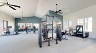 Located at 24720 Morton Ranch Rd in Katy, TX, this well-appointed fitness center offers a generous, single-floor layout designed to support a full range of workout routines. The space is architecturally distinguished by soaring vaulted ceilings accented with a distinctive teal vertical shiplap feature wall, lending a modern yet approachable aesthetic. Large windows on multiple sides flood the interior with natural light and provide views of the surrounding neighborhood, while multiple ceiling fans ensure comfortable air circulation throughout.

The facility is divided into clearly defined zones for both strength training and cardio. The strength area is equipped with multi-station cable machines, free-weight benches, weight racks, and functional training rigs including a TRX suspension system and a heavy bag. The cardio zone features interactive spin bikes, rowing machines, treadmills, stair climbers, and stationary bikes, several outfitted with personal display screens. Full-length mirrors along one wall enhance the sense of space and support proper form during workouts.

Entry is provided through sleek black-framed glass double doors, reinforcing the facility's contemporary design language. The light-toned flooring and crisp white walls create a clean, energizing environment well-suited for daily fitness activity.