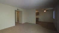 This single-floor residential unit offers a straightforward, functional layout designed for comfortable everyday living. The main living area is spacious and open, finished with wall-to-wall carpeting in a neutral beige tone that adds warmth underfoot. The walls are painted in a light, neutral color, contributing to a bright and airy atmosphere throughout.

A ceiling fan is positioned in the adjoining dining or secondary living area, providing both air circulation and overhead lighting. This area connects seamlessly to the kitchen, which features dark wood-toned cabinetry offering ample storage space. The kitchen is equipped with essential appliances and provides a functional workspace for meal preparation.

A textured ceiling runs throughout the unit, a common architectural detail that adds subtle visual interest. A hallway visible from the main living area leads to additional rooms, including what appears to be a bathroom and at least one other room, suggesting a multi-room floor plan. HVAC vents are integrated into the ceiling, ensuring climate control throughout the space.

Overall, the unit presents a clean, modest, and well-organized living environment with a neutral palette that offers flexibility for personalization and furnishing.