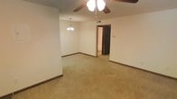 This single-floor residence offers a straightforward and functional layout designed for comfortable everyday living. The main living area is finished with wall-to-wall carpeting in a neutral tone, providing a warm and cohesive foundation throughout the space. White walls and dark wood baseboards create a clean contrast that keeps the interior feeling bright and tidy. A ceiling fan with an integrated light fixture is centrally positioned overhead, ensuring both air circulation and ambient lighting. An elegant chandelier-style pendant light adds a classic decorative touch, distinguishing the dining or transitional area from the main living zone. The open layout allows for flexible furniture arrangements, making the space adaptable to a variety of needs. A doorway visible from the main area leads to an additional room, suggesting a connected and navigable floor plan that flows naturally between spaces. The overall ambiance is understated and neutral, providing a versatile canvas well-suited for a range of interior styles. The single-floor design ensures ease of accessibility, and the consistent finishes throughout lend the space a unified, cohesive character.