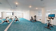 Located in Winter Garden, Florida, this expansive two-story fitness center offers a comprehensive and well-appointed wellness facility. The main floor is predominantly dedicated to cardio and strength training, featuring vibrant teal carpet flooring throughout and an abundance of natural light streaming in through large floor-to-ceiling windows that frame views of palm trees and the surrounding outdoor amenities, including a pool. Rows of treadmills, ellipticals, rowing machines, stair climbers, and stationary bikes are thoughtfully arranged across the spacious open floor plan, complemented by floor-to-ceiling mirrors and a bold geometric accent wall with built-in storage cubbies. A dedicated cycling studio houses multiple spin bikes and a wall-mounted display screen, while a separate yoga and barre room features elegant herringbone hardwood flooring, mirrored walls, barre rails, yoga mats, and large windows with outdoor views. An adjacent multi-purpose room, also finished with herringbone wood floors and textured blue-grey wall coverings, serves as a golf and game area equipped with putting equipment and a wall-mounted television. Recessed lighting throughout both floors ensures a bright, modern ambiance, and open-railing staircases connect the two levels seamlessly, creating a cohesive and energizing environment designed to support a full range of fitness activities.
