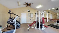 This single-story fitness room offers a functional and comfortable workout environment. The space features vaulted ceilings that create an open, airy atmosphere, complemented by a ceiling fan that aids in air circulation during exercise sessions. The walls are painted in a warm, neutral tone, contributing to an inviting ambiance. The room is carpeted throughout, providing a softer surface underfoot.

The fitness area is equipped with a multi-station cable weight machine with red upholstered benches, suitable for a variety of strength-training exercises. An elliptical trainer is also present, offering cardiovascular workout options. A large wall mirror on one side enhances the sense of space and allows users to monitor their form. A wall clock and several informational charts or notices are mounted on the walls, adding practical utility to the space.

Lighting is provided by a fluorescent ceiling fixture, ensuring adequate illumination throughout the room. A single door provides access, keeping the space well-defined and private. Overall, the room is designed to support a well-rounded fitness routine in a straightforward, no-frills setting.