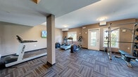 This exercise room is a comprehensively outfitted fitness facility contained on a single level, offering a generous open floor plan designed to accommodate a wide variety of workout activities. The space is finished with durable, patterned carpet tile in muted grey and brown tones, providing both traction and a clean aesthetic throughout.

The cardio zone features multiple treadmills, an elliptical trainer, and a rowing machine, positioned near windows that allow natural light to fill the space and offer views of the surrounding outdoor environment. A door provides direct exterior access, further enhancing ventilation and connectivity to the outdoors.

The strength training area is equally well-appointed, including a multi-function cable machine, an adjustable weight bench, and a full dumbbell rack. Full-length mirrors line one wall, visually expanding the space and supporting proper form during exercise. Wall-mounted flat-screen televisions are positioned throughout for entertainment during workouts.

Structurally, the room features support columns and a drop soffit detail that define distinct zones within the open layout. Climate control is provided by a wall-mounted split unit, ensuring year-round comfort. The entrance area includes accessible washroom facilities and a glass-paneled entry door. Flush-mount ceiling lighting provides consistent illumination throughout the space.