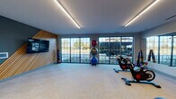 Located at 2100 Progress Pkwy in Schaumburg, Illinois, this single-floor fitness facility offers a thoughtfully designed workout environment that blends contemporary aesthetics with functional equipment. The main exercise area is defined by large, floor-to-ceiling grid-pane windows along one wall, flooding the space with natural light and providing open views of the surrounding landscape. Linear LED lighting strips run across the smooth ceiling, ensuring consistent illumination throughout.

A striking accent wall features diagonally arranged wood slat paneling set against a dark charcoal background, creating a bold visual focal point. Mounted on this wall is a large flat-screen display, ideal for guided workouts or informational content. Neutral-toned carpet tile flooring extends throughout, providing a clean, low-maintenance surface suitable for a variety of exercises.

The facility is well-stocked with spinning bikes, kettlebells, medicine balls, stability balls, a TRX suspension system with overhead mounting bars, and a multi-functional cable machine with dumbbell storage. Built-in lockers offer convenient storage for users. Additional rooms accessible via wood-panel doors provide private changing or restroom facilities. Overall, the space delivers a professional, modern fitness experience.
