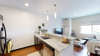 This single-floor unit offers a thoughtfully designed open-concept layout that seamlessly connects the kitchen and living area. The kitchen is a standout feature, boasting granite countertops with distinctive veining, dark espresso cabinetry, and a peninsula with bar seating that doubles as a natural divider between the kitchen and living space. Pendant lighting above the peninsula adds a stylish touch, while black appliances — including a full-size refrigerator, range, and built-in microwave — provide a sleek, cohesive look. Warm hardwood flooring flows throughout the unit, lending a sense of continuity and comfort.

The living area is open and inviting, receiving natural light through a large window that offers views to the outdoors. The space comfortably accommodates a seating area and entertainment setup, with the open sightlines to the kitchen enhancing the overall sense of space.

The bathroom continues the granite and dark cabinetry theme found in the kitchen, featuring a single undermount sink, a large wall mirror with overhead lighting, and a shower-tub combination. Neutral wall tones throughout the unit create a clean, versatile backdrop. Overall, the space presents a modern and functional design with consistent finishes that give it a cohesive and polished character.