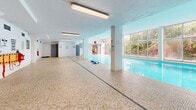 Located at 50 Hillsboro Ave in Toronto's sought-after Annex neighbourhood, this generously proportioned indoor recreational facility offers a compelling combination of aquatic and fitness amenities across a single expansive floor. The centrepiece of the space is a full-length indoor swimming pool, its vivid turquoise water set against warm mosaic tile surrounds that extend across the pool deck. Floor-to-ceiling windows run along one side of the pool area, flooding the interior with natural light and framing views of mature, leafy greenery outside — creating a bright and refreshing atmosphere year-round. Entry to the pool deck is accessible via a glass-panelled door that opens directly to an outdoor terrace, further connecting the interior to the natural surroundings. Structural columns punctuate the pool perimeter, complementing the clean, institutional architecture of the space. Safety equipment, including life rings and clearly marked signage, is thoughtfully positioned throughout. Adjacent to the pool area, a dedicated fitness zone houses cardio and strength training equipment, including a treadmill, elliptical, weight bench, and pull-up station, alongside wall-mounted mirrors to support a complete workout. The facility also includes change room access and additional utility areas, making it a well-rounded recreational amenity.