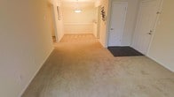 This single-floor space offers a clean, open layout finished in warm neutral tones throughout. The main living area is carpeted in a soft beige, creating a comfortable and cohesive feel underfoot. A pendant light fixture hangs centrally, providing overhead illumination and serving as a subtle focal point in the open room. The walls are painted in a light cream tone, contributing to the bright and airy ambiance of the interior. A chair rail detail runs along the wall, adding a classic architectural touch to the space. Toward the entry, the flooring transitions from carpet to a dark hardwood or laminate surface, delineating the entryway from the main living zone. A white door with standard hardware provides access to the exterior or an adjacent area, while wall-mounted coat or accessory racks nearby offer practical storage near the entrance. An additional doorway or opening at the far end of the space suggests a connected hallway or adjoining room, allowing for easy flow throughout the floor plan. A framed piece of wall art adds a personal touch to the otherwise minimal interior. Overall, the space presents a straightforward, functional design with a warm and inviting atmosphere.