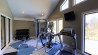 This fitness center is thoughtfully designed to support a comprehensive workout experience. The space features soaring vaulted ceilings that create an open, airy atmosphere, complemented by large windows positioned high along the walls, allowing abundant natural light to pour in while providing views of the surrounding outdoors. The neutral, modern color palette of gray tones and white trim gives the room a clean, professional appearance. The flooring consists of a patterned area rug over hard flooring, providing both comfort and durability underfoot. A wall-mounted flat-screen television is conveniently positioned for entertainment during workouts. The equipment layout is well-organized, featuring a multi-station cable machine for strength training, an elliptical and a treadmill with integrated display screens for cardio workouts, and a dedicated free weights area with a full rack of dumbbells and an adjustable bench. Recessed lighting supplements the natural light, ensuring the space remains bright and functional at any time of day. The overall design balances practicality with a welcoming ambiance, making this fitness center a versatile and motivating environment for a wide range of exercise routines.