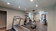 This dedicated fitness center offers a comprehensive and well-organized workout environment designed to support a full range of exercise routines. The space is finished with neutral gray walls and durable rubber flooring, creating a practical and clean aesthetic throughout. Recessed lighting illuminates the room evenly, contributing to a bright and energizing atmosphere.

The cardio section is equipped with a treadmill, an elliptical trainer, and a stationary cycle, providing a solid variety of aerobic exercise options. A large wall mirror spans one side of the space, visually expanding the room and allowing users to monitor their form during workouts. A multi-station cable and weight machine anchors the strength training area, complemented by a rowing-style machine and a barbell, offering versatile resistance training capabilities. Stability balls of varying sizes are available for core and flexibility exercises.

Natural light filters in through a window that looks out onto lush greenery, adding a refreshing connection to the outdoors. The layout is thoughtfully arranged to maximize usable floor space while keeping equipment accessible. Safety features, including a mounted fire extinguisher, are clearly visible. This fitness center provides a well-rounded facility suitable for daily exercise needs.