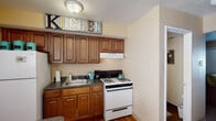 This two-floor home offers a practical and comfortable layout across its living spaces. The kitchen is equipped with rich warm-toned wood cabinetry, dark granite-style countertops, a gas range, and a full-size refrigerator, all set over hardwood flooring that adds warmth to the space. Adjacent to the kitchen, the dining area features hardwood floors and a dark wood dining table beneath decorative wall art, with a louvered storage closet nearby. The living room is carpeted and provides a cozy gathering space with a front-facing window that allows natural light to filter in. Upstairs, the primary bedroom is a serene retreat with carpeted floors, a sliding closet, natural light from a window with curtains and blinds, and tasteful wall art. A second bedroom is designed with a child in mind, featuring carpet flooring, a bright window, and a charming, playful aesthetic. A hallway with a staircase railing connects the upper-floor rooms, with a wooden dresser and mirror visible from the primary bedroom doorway. Throughout the home, textured ceilings and neutral wall tones create a consistent, understated backdrop across all rooms.