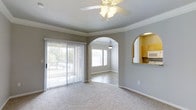 This single-story, one-bedroom, one-bathroom unit offers a thoughtfully arranged layout that maximizes comfort and functionality. The living area is anchored by plush carpet flooring and features a ceiling fan with a light fixture overhead, along with crown molding that adds a refined architectural touch. A sliding glass door fitted with vertical blinds allows natural light to pour in while providing access to outdoor views of surrounding greenery.

A distinctive arched passageway connects the living area to a secondary space — likely a dining area — which features hardwood-style flooring, a hanging pendant light, and a bright window that further enhances the natural light throughout. This arched design element adds character and visual depth to the open flow of the unit.

Adjacent to the living space, a pass-through kitchen window with a rounded arch opening reveals a kitchen outfitted with wooden cabinetry and stainless steel appliances, including a built-in microwave. The neutral gray wall tones throughout the unit create a calm, modern ambiance, while the combination of carpet and hard-surface flooring adds variety and practicality to the overall design.