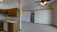 This single-floor space features an open-concept layout that connects the kitchen and living areas seamlessly. The living area is finished with neutral-toned carpet, creating a warm and understated foundation throughout the space. White walls, accented by dark wood baseboards and crown molding, provide a clean contrast that adds visual definition to each room.

A ceiling fan with a light fixture is centrally positioned overhead, contributing both to air circulation and ambient lighting. The kitchen area, partially visible to one side, includes warm honey-toned wood cabinetry with traditional raised-panel doors, offering ample storage. A white countertop and a structural support column are also present near the kitchen, subtly separating it from the main living space.

A hallway visible in the background leads to additional rooms, with a door at the far end suggesting access to a bathroom or bedroom. A floor vent indicates a forced-air HVAC system is in place. The overall ambiance of the space is practical and move-in ready, with a traditional aesthetic that emphasizes functionality and comfort throughout.