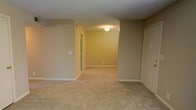 This single-floor space presents a straightforward and functional layout with a clean, neutral aesthetic throughout. The living area is finished with wall-to-wall beige carpeting that adds warmth underfoot and contributes to the overall subdued color palette. Walls are painted in a soft tan hue, complemented by crisp white baseboards that define the perimeter of each room.

The textured popcorn ceiling adds a classic touch and houses a centrally mounted flush light fixture that provides ambient illumination to the space. An open passageway connects the main living area to an adjoining room, creating a sense of continuity and flow between the two spaces. This secondary room mirrors the same carpeting and wall finishes, maintaining a cohesive look throughout.

A doorway on the right side of the main area leads to an additional room, while another opening on the left side provides access to a further space or hallway, suggesting a multi-room configuration. Electrical outlets and switch plates are positioned at standard heights along the walls. The overall atmosphere is understated and move-in ready, offering a blank canvas well-suited for a variety of furniture arrangements and personal styling choices.
