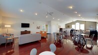 This single-floor community space offers an open-concept layout that combines a comfortable lounge area with a casual dining and gathering space. The room is defined by striking vaulted ceilings that rise dramatically overhead, enhancing the sense of spaciousness and airiness throughout. Recessed lighting and ceiling fans are thoughtfully distributed across the ceiling, ensuring both comfort and illumination throughout the space.

The rich, high-gloss hardwood flooring runs the length of the room, adding warmth and a polished aesthetic that contrasts elegantly with the crisp white walls and ceiling. The lounge area features a neutral gray sofa and accent chairs in bold orange, arranged around a wall-mounted flat-screen television atop a media console, creating an inviting spot for relaxation.

The dining area is furnished with multiple round tables paired with modern industrial-style metal chairs, offering flexible seating arrangements for gatherings. Natural light filters in through windows along the far wall, brightening the space further. A second ceiling fan is visible toward the rear of the room, reinforcing the generous scale of the layout. Access doors are positioned at the back, providing convenient connectivity to adjacent areas. The overall ambiance is clean, modern, and welcoming.