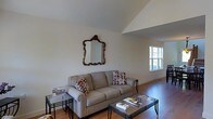 This two-story space offers a thoughtfully designed interior with notable architectural details. The living area is a standout feature, showcasing soaring vaulted ceilings that lend an open, airy quality to the room. The ceiling's angled lines add visual interest and a sense of generous vertical space.

Hardwood flooring runs through the living area, providing warmth and a classic aesthetic that complements the neutral color palette of the walls. Natural light filters in through a multi-pane window, brightening the space and creating a welcoming ambiance.

The living area is centered around a comfortable taupe sofa accented with patterned throw pillows in warm earth tones. An ornate gold-framed mirror serves as a focal wall accent, adding a touch of elegance. Glass-top coffee and side tables with wrought iron frames complete the seating arrangement, balancing the traditional and contemporary elements of the room.

The overall design is clean and understated, with neutral tones throughout creating a cohesive and calm environment. The combination of vaulted ceilings, natural light, and hardwood floors makes this a particularly inviting living space.
