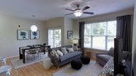 This single-floor space seamlessly integrates a living area and dining space within an open-plan layout. The living area is anchored by a gray upholstered sofa adorned with an assortment of decorative pillows, complemented by a patterned area rug with a sunburst motif in muted yellows and grays. A ceiling fan with wooden blades serves as both a functional and aesthetic centerpiece overhead. Two knitted black poufs and a white ceramic garden stool add versatile seating options throughout the space. Natural light floods the room through multiple windows fitted with horizontal blinds and dark curtain panels, as well as a sliding glass door that opens to an outdoor area, offering views of lush greenery. The dining area, positioned adjacent to the living space, features a wooden table set with dark chairs and is illuminated by a distinctive white globe pendant light. Light hardwood flooring runs throughout, enhancing the bright and airy ambiance. Wall art and a table lamp add finishing touches, contributing to the warm, inviting character of the space.