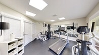 This fitness center at 100 Bent Tree Dr in Daytona Beach, Florida, is a single-room facility designed to provide residents with a convenient and functional workout space. The room features durable dark rubber flooring, neutral light-colored walls, and a bright, clean aesthetic throughout. Illumination is provided by recessed flat-panel ceiling lights that evenly distribute light across the space, creating a well-lit environment conducive to exercise. A large wall-mounted mirror spans one side of the room, effectively amplifying the sense of space and allowing users to monitor their form during workouts. The fitness center is equipped with a range of machines and equipment, including a Landice treadmill, an elliptical cross-trainer, a multi-station cable strength machine with attachments for various exercises, and a set of free weights and dumbbells. A wall-mounted flat-screen television is positioned for easy viewing during workouts. A large window on one wall allows natural light to enter and offers a view of the surrounding greenery outside. Louvered shutter doors provide access to storage or adjacent areas. A small shelving unit offers additional organizational space. The room is well-ventilated with visible HVAC vents, and exercise room rules are posted on the wall, indicating this is a shared community amenity.