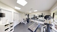This fitness center at 100 Bent Tree Dr in Daytona Beach, Florida, is a single-room facility designed to provide residents with a convenient and functional workout space. The room features durable dark rubber flooring, neutral light-colored walls, and a bright, clean aesthetic throughout. Illumination is provided by recessed flat-panel ceiling lights that evenly distribute light across the space, creating a well-lit environment conducive to exercise. A large wall-mounted mirror spans one side of the room, effectively amplifying the sense of space and allowing users to monitor their form during workouts. The fitness center is equipped with a range of machines and equipment, including a Landice treadmill, an elliptical cross-trainer, a multi-station cable strength machine with attachments for various exercises, and a set of free weights and dumbbells. A wall-mounted flat-screen television is positioned for easy viewing during workouts. A large window on one wall allows natural light to enter and offers a view of the surrounding greenery outside. Louvered shutter doors provide access to storage or adjacent areas. A small shelving unit offers additional organizational space. The room is well-ventilated with visible HVAC vents, and exercise room rules are posted on the wall, indicating this is a shared community amenity.