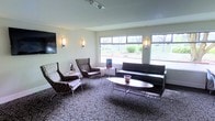 This single-floor space presents a thoughtfully designed lounge or reception area that blends contemporary style with functional comfort. The room is anchored by large picture windows that flood the interior with natural light while offering pleasant views of the surrounding greenery and landscaping outside.

The seating arrangement is both varied and inviting, featuring a sleek black leather sofa paired with a modern oval coffee table, complemented by a set of patterned accent chairs with a classic mid-century aesthetic. This mix of furniture styles gives the space a layered, curated character that balances sophistication with approachability.

Underfoot, a boldly patterned carpet with an interlocking circular motif adds visual interest and depth to the floor, tying the room's design elements together. Wall-mounted sconce lighting provides warm, ambient illumination throughout, supplementing the natural light from the windows. A flat-screen television is mounted on one wall, adding a practical, modern touch to the space.

The walls are finished in a clean, neutral tone that keeps the atmosphere open and airy, allowing the furnishings and décor to take center stage. Overall, this space exudes a professional yet welcoming ambiance, well-suited for hosting guests, clients, or visitors in a relaxed and comfortable setting.