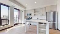 This thoughtfully designed single-floor residence showcases a clean, contemporary aesthetic with a cohesive neutral palette throughout. The heart of the home is an open-concept kitchen, where light gray cabinetry with matte black hardware pairs elegantly with a white subway tile backsplash and light hardwood-style flooring. A central island with a white quartz countertop provides both additional prep space and built-in open shelving, ideal for storage and display. Stainless steel appliances — including a gas range, over-the-range microwave, and French door refrigerator — lend a polished, functional character to the space. A large window near the kitchen floods the area with natural light, offering views of the surrounding exterior. The bathroom continues the modern design language, featuring white cabinetry with matching black hardware, a full tub-shower combination with white tile surround, and wood-style flooring that extends from the main living area. A dedicated bedroom offers a quieter, cozier atmosphere with plush carpeting, a wall-mounted accent light, and textured wall panels that add warmth and dimension. Recessed lighting throughout ensures the space remains bright and well-illuminated, while dark-framed doors provide striking contrast against the bright, airy interiors.