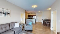 This single-floor unit offers an open-concept layout that seamlessly connects the living, dining, and kitchen areas into one cohesive, functional space. The living area is anchored by a plush gray sofa and armchair accented with teal throw pillows, complemented by a small side table and lamp that add a warm, inviting touch. Light-toned, hardwood-style flooring extends throughout, enhancing the sense of openness and continuity.

The kitchen features rich, dark wood cabinetry paired with dark countertops, creating a striking contrast against the lighter surroundings. Stainless steel appliances, including a refrigerator and microwave, add a modern finish, while a peninsula provides additional prep and seating space. Pendant lighting above the kitchen area adds both style and focused illumination.

Adjacent to the kitchen, a compact dining area accommodates a dark wood table and chairs, ideal for everyday meals. Doorways visible throughout the space suggest additional rooms nearby. Neutral gray walls and flush ceiling lighting contribute to a clean, contemporary ambiance, while the open floor plan ensures excellent flow and versatility throughout the unit.