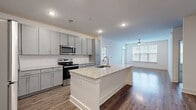 This single-floor, 2-bedroom, 2-bathroom residence offers a thoughtfully designed open-concept layout that maximizes functionality and flow. The kitchen serves as the centerpiece of the living area, showcasing a large central island with a granite countertop and undermount sink, grey shaker-style cabinetry extending to the ceiling, a white subway tile backsplash, and a full suite of stainless steel appliances including a refrigerator, range, and microwave. The kitchen opens seamlessly into the living room, which is bathed in natural light from large windows and features a ceiling fan and wood-look flooring that runs throughout the main living areas. Both bedrooms are carpeted and offer a clean, neutral palette with ample wall space. The spacious primary bedroom includes its own dedicated closet with wire shelving for organized storage. A second closet with similar wire shelving serves the additional bedroom. Each bathroom is finished with granite-topped grey vanities, large mirrors, wood-look flooring, and a soaking tub with shower combination. The consistent use of grey tones, granite surfaces, and modern hardware creates a cohesive and contemporary aesthetic throughout the entire residence.