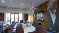 This single-floor space is designed with a contemporary aesthetic, combining a lounge area and a formal meeting space within an open, well-organized layout. The lounge area features dark leather seating arranged around layered stone coffee tables, all set atop a rich, patterned red carpet that adds depth and visual interest to the room. A striking large-scale portrait is displayed on a textured wood-panel accent wall, which also houses multiple mounted flat-screen televisions at varying heights, creating a dynamic media focal point. Recessed lighting throughout the ceiling provides consistent, ambient illumination, complementing the clean white walls and enhancing the bold design contrasts. A bold red abstract artwork adds a further artistic element to the space. Large glass doors along one wall allow natural light to filter in and offer views to the exterior. Adjacent to the lounge, a formal conference or meeting area is defined by a deep-toned table surrounded by navy blue chairs, offering a professional setting suitable for meetings or collaborative work. The overall ambiance is polished and modern, blending functional workspace with a stylish, hospitality-inspired atmosphere.