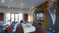 This single-floor space is designed with a contemporary aesthetic, combining a lounge area and a formal meeting space within an open, well-organized layout. The lounge area features dark leather seating arranged around layered stone coffee tables, all set atop a rich, patterned red carpet that adds depth and visual interest to the room. A striking large-scale portrait is displayed on a textured wood-panel accent wall, which also houses multiple mounted flat-screen televisions at varying heights, creating a dynamic media focal point. Recessed lighting throughout the ceiling provides consistent, ambient illumination, complementing the clean white walls and enhancing the bold design contrasts. A bold red abstract artwork adds a further artistic element to the space. Large glass doors along one wall allow natural light to filter in and offer views to the exterior. Adjacent to the lounge, a formal conference or meeting area is defined by a deep-toned table surrounded by navy blue chairs, offering a professional setting suitable for meetings or collaborative work. The overall ambiance is polished and modern, blending functional workspace with a stylish, hospitality-inspired atmosphere.