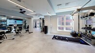 Located at 95 Chestnut St in Providence, Rhode Island, this well-appointed fitness center offers a comprehensive workout environment spread across a single, thoughtfully organized floor. The space is designed with a clean, modern aesthetic, featuring crisp white walls, light-toned flooring, and recessed linear LED lighting that creates a bright, energizing atmosphere throughout.

The facility is divided into distinct functional zones. A dedicated cardio area lines one side of the space, outfitted with treadmills, StairMasters, and elliptical machines positioned in front of large windows that frame views of the surrounding Providence cityscape, allowing natural light to pour in during workouts. Ceiling fans are mounted overhead for added comfort.

The strength training area is generously equipped with a full rack of dumbbells, adjustable benches, cable machines, and a power rack system. This section is anchored by a striking large-scale mural featuring illustrated fitness motifs — bicycles, kettlebells, and dynamic line work — rendered in red and white, adding an energetic focal point to the room.

A separate, open functional training zone provides ample floor space for stretching, bodyweight exercises, and mat-based workouts, accompanied by wall-mounted screens. Mirrored walls throughout enhance the sense of space and support form-checking during training. The facility's layout ensures a smooth, unobstructed flow between all workout areas.