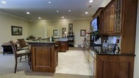 This single-floor space exudes a warm and polished ambiance, blending classic design elements with practical functionality. The interior is anchored by rich, medium-toned wood cabinetry with raised-panel detailing, complemented by sleek dark granite countertops that run throughout. The flooring consists of large-format, light-colored travertine-style tiles that lend an air of sophistication and openness to the space.

Recessed lighting is evenly distributed across the ceiling, providing consistent and well-balanced illumination throughout. Crown molding adds a refined architectural touch, framing the room with a traditional yet timeless character. A central island or service counter serves as a focal point, offering additional workspace and a natural gathering area within the layout.

Along one wall, a built-in cabinetry unit houses a large flat-screen television, creating a multimedia or lounge-oriented element within the space. A coffee or beverage station is also present, complete with cups and accessories, suggesting a hospitality or communal use. Decorative wall art and a warm table lamp add character and a lived-in comfort to the environment. The open layout and thoughtful design make this space feel both inviting and highly functional.
