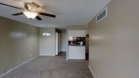 This single-floor residence presents a practical, open-concept layout that connects the living and dining areas in one cohesive space. The living area is finished with wall-to-wall carpet in a neutral tan tone, complemented by warm beige walls that lend a consistent, understated palette throughout. A ceiling fan with a light fixture is positioned in the living area, while a decorative chandelier marks the dining space, adding a subtle touch of elegance. The textured popcorn ceiling runs throughout, adding a classic residential character.

The kitchen is positioned at the rear of the open floor plan and features a raised breakfast bar-style pass-through that opens toward the dining and living areas, promoting an interconnected feel. Dark stainless appliances, including a microwave and refrigerator, are visible within the kitchen, which transitions to hard flooring — contrasting with the carpeted living spaces. Adjacent to the kitchen is a built-in storage area with louvered bi-fold doors and additional cabinetry, providing ample organizational space. A hallway connects to further rooms, completing the functional flow of this well-organized single-level layout.