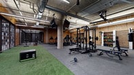 Located at 230 East Ave in Norwalk, Connecticut, this fitness center combines an industrial aesthetic with a comprehensive range of workout amenities. The main training floor is expansive and open, featuring exposed concrete ceilings with visible ductwork and structural columns that contribute to the space's raw, urban character. Oriented strand board (OSB) wall paneling reinforces the industrial design throughout.

The primary workout area is thoughtfully zoned to accommodate a variety of training styles. A dedicated strength section houses multiple power racks, barbells, weight plates, and adjustable benches, while a row of treadmills and elliptical machines lines the perimeter for cardio training. A turf strip running through the center of the floor provides space for functional fitness drills and sled pushes. Mounted flat-screen monitors are positioned throughout for entertainment or instructional use.

A separate, enclosed spin studio offers a focused environment for cycling classes, equipped with a fleet of stationary bikes and built-in shelving, set against dark-painted walls for an immersive atmosphere. An additional glass-partitioned room serves as an equipment storage and amenity area, featuring organized shelving and a clean, accessible layout. The facility is well-lit with linear fluorescent and LED fixtures, creating a bright and functional training environment throughout.