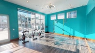 This well-appointed fitness studio is designed to energize and motivate, featuring bold teal accent walls that create a dynamic, high-energy atmosphere throughout the space. The room is anchored by a row of stationary spin bikes positioned directly in front of expansive floor-to-ceiling windows, flooding the workout area with abundant natural light and offering unobstructed views of an outdoor courtyard and surrounding building community.

The flooring throughout consists of durable wood-style planks with warm-toned baseboards, providing a clean and modern foundation. A large wall mirror on one end visually expands the space, while a wall-mounted flat-screen television offers entertainment during workouts. Recessed lighting and a circular ceiling fixture provide consistent, even illumination throughout.

Along the teal accent wall, wall-mounted TRX suspension training straps and resistance-based equipment offer versatile bodyweight training options. Built-in wooden storage cubbies and shelving units line portions of the walls, providing organized storage for accessories and fitness supplies. Upper windows along secondary walls admit additional natural light.

The studio's single-level layout is open and unobstructed, making it well-suited for group fitness classes or individual training sessions. The overall ambiance is bright, modern, and inviting.