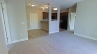 This single-floor residence features an open-concept layout that seamlessly connects the living, dining, and kitchen areas. The living area is finished with neutral-toned carpet, providing a comfortable and cohesive feel, while the kitchen and adjoining dining space transition to tile flooring. A partial wall with a pass-through counter separates the kitchen from the living area, maintaining an open and airy atmosphere while defining each functional zone.

The kitchen is equipped with dark espresso-toned cabinetry and black appliances, including a full-size refrigerator with an integrated dispenser. Flush-mount ceiling lighting illuminates both the kitchen and living areas, contributing to a well-lit interior. A built-in desk or bar-height counter nook is tucked along one wall near the entryway, offering a practical workspace or additional surface area.

The entryway features a classic six-panel door and is finished with the same neutral wall tones found throughout the space. A doorway visible from the main living area leads to an additional room, suggesting a versatile and functional floor plan. The overall aesthetic is clean, neutral, and move-in ready.