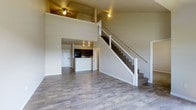 This two-story space showcases an open and airy design that immediately impresses with its soaring double-height ceilings in the main living area. Wide-plank, hardwood-style flooring in a warm gray tone extends throughout the main level, lending a cohesive and modern feel to the space. The expansive living area is flooded with a sense of openness, enhanced by the dramatic vertical space above and a ceiling fan that adds both function and character.

A beautifully crafted staircase with white-painted wooden balusters and dark metal spindles serves as a striking architectural focal point, leading gracefully to the upper level. The open railing on the second floor overlooks the living area below, reinforcing the connected, two-story feel.

Adjacent to the living area, the kitchen is thoughtfully integrated into the layout, featuring dark cabinetry, a central island or peninsula, and track lighting overhead. Black appliances add a bold contrast to the lighter surroundings. A doorway near the kitchen provides convenient access to an entry or utility area, completing the practical flow of the main floor.

The neutral color palette of warm whites and soft beiges throughout creates a clean, inviting ambiance that allows the architectural details and layout to take center stage.