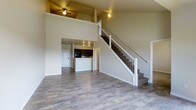 This two-story space showcases an open and airy design that immediately impresses with its soaring double-height ceilings in the main living area. Wide-plank, hardwood-style flooring in a warm gray tone extends throughout the main level, lending a cohesive and modern feel to the space. The expansive living area is flooded with a sense of openness, enhanced by the dramatic vertical space above and a ceiling fan that adds both function and character.

A beautifully crafted staircase with white-painted wooden balusters and dark metal spindles serves as a striking architectural focal point, leading gracefully to the upper level. The open railing on the second floor overlooks the living area below, reinforcing the connected, two-story feel.

Adjacent to the living area, the kitchen is thoughtfully integrated into the layout, featuring dark cabinetry, a central island or peninsula, and track lighting overhead. Black appliances add a bold contrast to the lighter surroundings. A doorway near the kitchen provides convenient access to an entry or utility area, completing the practical flow of the main floor.

The neutral color palette of warm whites and soft beiges throughout creates a clean, inviting ambiance that allows the architectural details and layout to take center stage.