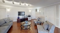 This single-floor communal space offers an open, well-connected layout that seamlessly integrates a living area, dining nook, and kitchen, making it a versatile and inviting environment. Rich hardwood flooring flows throughout the entire space, complemented by crisp white crown molding and chair-rail wainscoting that lend a refined, classic character to every room.

The living area is generously sized, featuring track lighting on the ceiling and wall sconces that provide warm, layered illumination. A media console with a wall-mounted flat-screen television anchors one side of the room, while comfortable seating arrangements with patterned accent chairs and a round glass coffee table encourage relaxed gathering. Paneled white doors and interior windows with grid detailing add architectural interest to the walls.

The dining nook is bright and airy, benefiting from a bank of large windows dressed with horizontal blinds that allow ample natural light to filter in while offering views of lush greenery outside. A round glass-top dining table with upholstered white chairs creates an intimate setting for meals or conversation.

The kitchen is practical and well-appointed, featuring warm wood cabinetry, laminate countertops, and a full suite of black appliances including a gas range, microwave, dishwasher, and refrigerator. A raised breakfast bar provides a casual divider between the kitchen and the main living space, enhancing the open, communal feel of the overall layout.