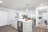 This well-appointed single-floor residence showcases a clean, contemporary design with an open-concept layout that connects the kitchen, dining, and living areas seamlessly. The kitchen serves as a central hub, featuring a large island with a white quartz countertop, bar seating, a stainless steel sink with a modern faucet, and stainless steel appliances including a refrigerator, electric range, and over-the-range microwave. White cabinetry with brushed nickel hardware and a subway tile backsplash complete the polished look. Pendant lighting illuminates the island, while recessed lighting brightens the entire open living space. Light wood-tone flooring flows throughout the main living areas, adding warmth and continuity. The dining area comfortably accommodates a full table and chairs, with direct sightlines to the kitchen. The living room benefits from multiple large windows fitted with horizontal blinds, allowing for generous natural light and views of surrounding greenery. Both bedrooms are carpeted and offer dedicated closets with built-in shelving. The bathrooms feature light wood cabinetry, white quartz countertops, undermount sinks, large mirrors, and wood-look flooring. One bathroom includes a shower, while the second serves as a half-bath style space. The overall aesthetic is bright, neutral, and modern throughout.