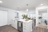 This well-appointed single-floor residence showcases a clean, contemporary design with an open-concept layout that connects the kitchen, dining, and living areas seamlessly. The kitchen serves as a central hub, featuring a large island with a white quartz countertop, bar seating, a stainless steel sink with a modern faucet, and stainless steel appliances including a refrigerator, electric range, and over-the-range microwave. White cabinetry with brushed nickel hardware and a subway tile backsplash complete the polished look. Pendant lighting illuminates the island, while recessed lighting brightens the entire open living space. Light wood-tone flooring flows throughout the main living areas, adding warmth and continuity. The dining area comfortably accommodates a full table and chairs, with direct sightlines to the kitchen. The living room benefits from multiple large windows fitted with horizontal blinds, allowing for generous natural light and views of surrounding greenery. Both bedrooms are carpeted and offer dedicated closets with built-in shelving. The bathrooms feature light wood cabinetry, white quartz countertops, undermount sinks, large mirrors, and wood-look flooring. One bathroom includes a shower, while the second serves as a half-bath style space. The overall aesthetic is bright, neutral, and modern throughout.